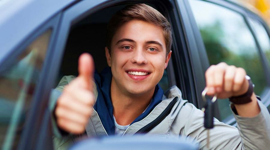 Young man smiling while sitting in car, giving thumbs up gesture and holding car keys out the window